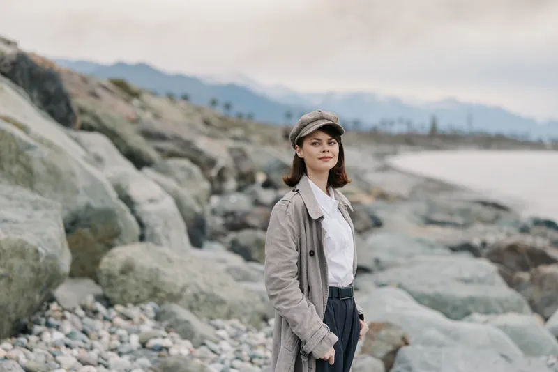 Young woman in a patterned coat and cap stands on a rocky beach with mountains in the background. Overcast sky creates a calm and contemplative mood outdoors.