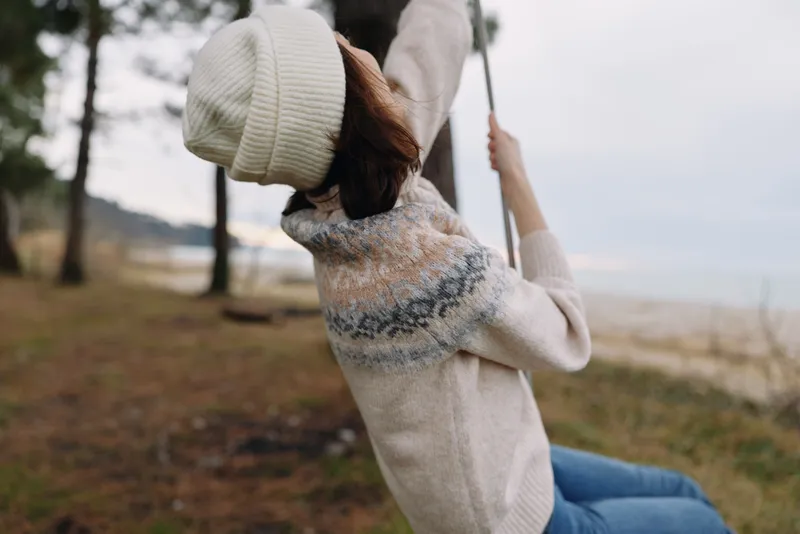 Outdoor swing moment with a woman in a knit sweater and beanie by the beach, autumn day, capturing carefree relaxation and motion today.