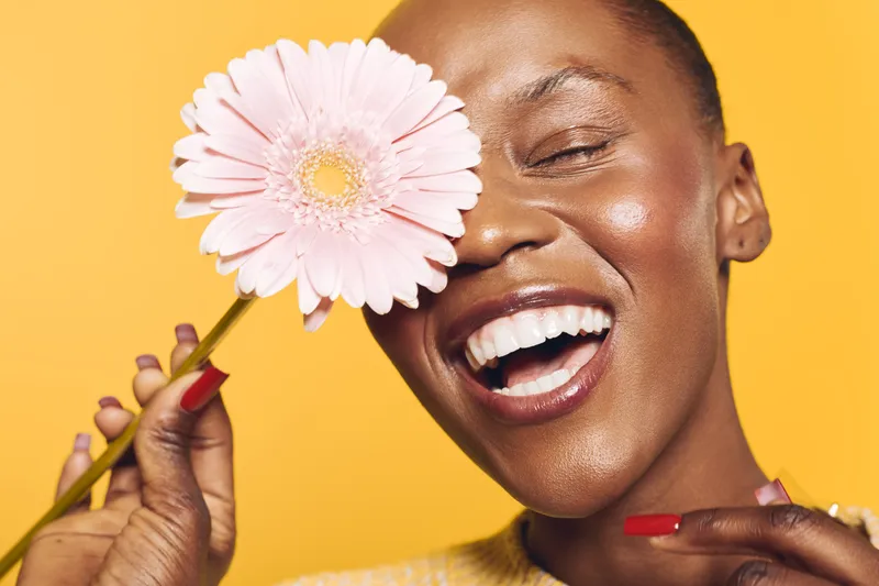 Happy African woman with dark skin holds pink flower near face smiling with closed eyes on yellow background. Concept of joy, beauty, natural skin, and floral wellness.