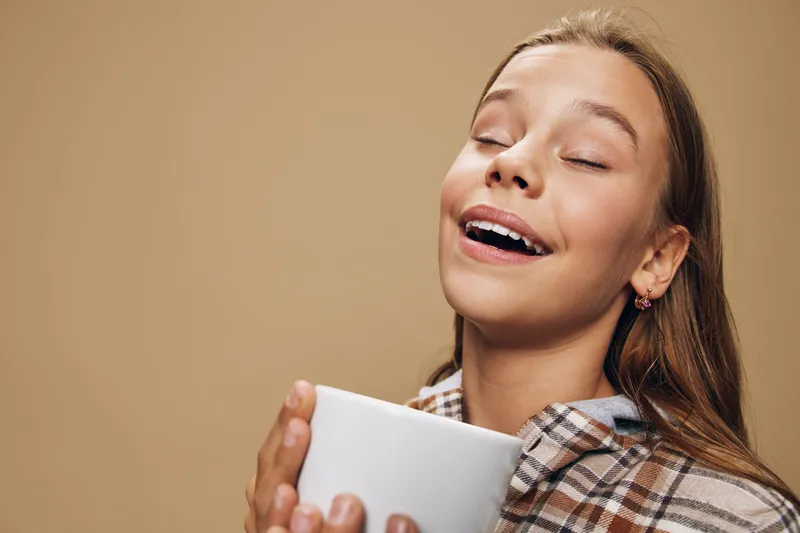 Young woman with closed eyes enjoying aroma of hot coffee or tea in hands wearing casual plaid shirt on beige background with peaceful expression and smile.