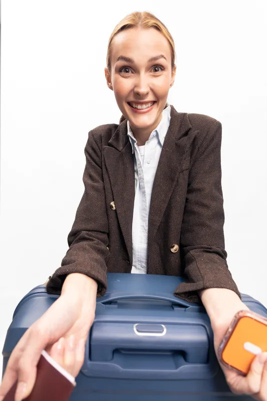 Smiling young woman in brown blazer holds passport and orange luggage tag over blue suitcase. Isolated on white background. Concept for travel, tourism, vacation and airport security.