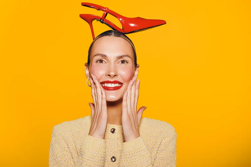 Smiling woman with red lipstick holds face with hands and wears red high heel shoes on head against bright yellow background. Concept of fashion, fun, and creative style.