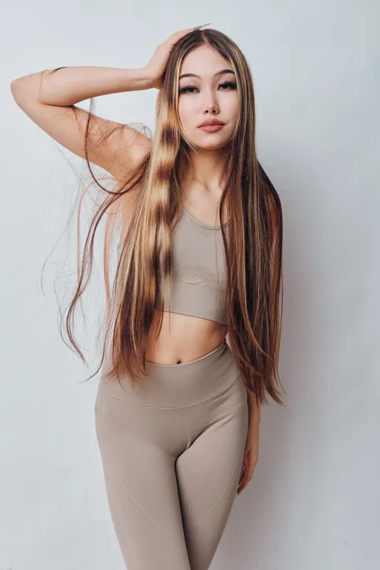 Young woman with long straight hair wearing sleeveless top and leggings poses against light background for fashion portrait, beauty, style and confident look indoors.