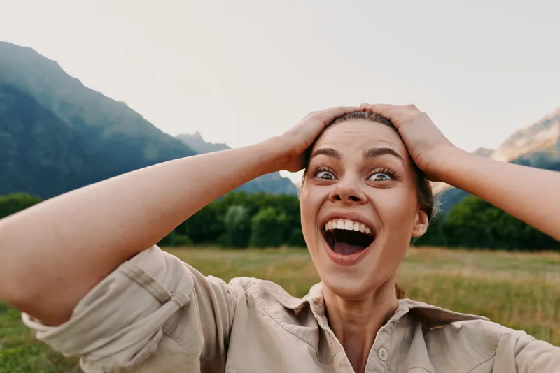 Young woman with short hair expresses surprise and joy while standing in a grassy field with mountains in the background on a clear day.