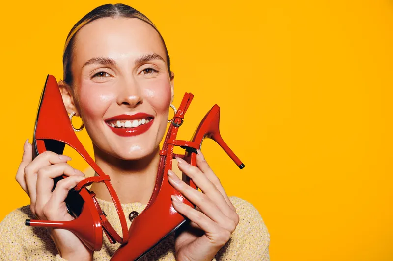 Smiling woman holds red high heel shoes near face on bright yellow background. Concept of fashion, style, happiness, and shopping with vibrant colors and joyful mood.