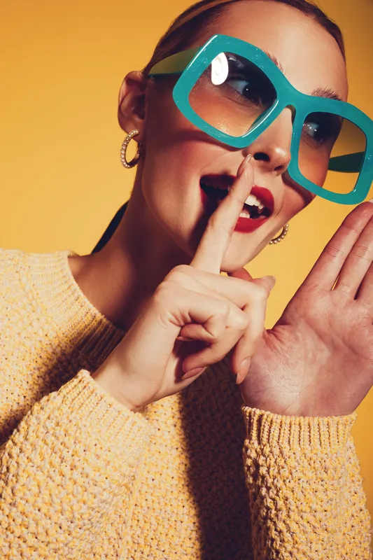 Young woman in oversized turquoise sunglasses and yellow sweater makes a shushing gesture with finger on lips. Bright studio lighting and yellow background create playful mood.