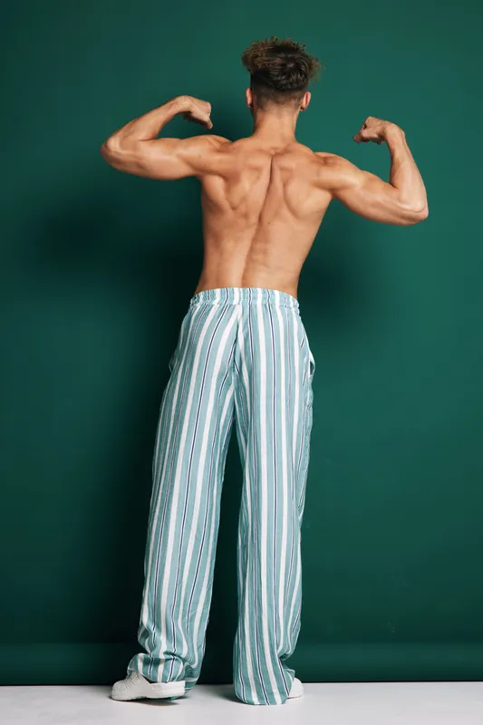 Fit young man showcasing muscular back and arms in loose striped pants posing against dark green background. Body, fitness, male model with defined muscles and curly hair indoors.
