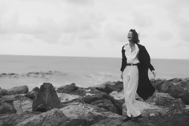 Monochrome scene of a woman walking along rocky coast, laughing, wearing a long coat and flowing pants, wind in her hair, seascape behind