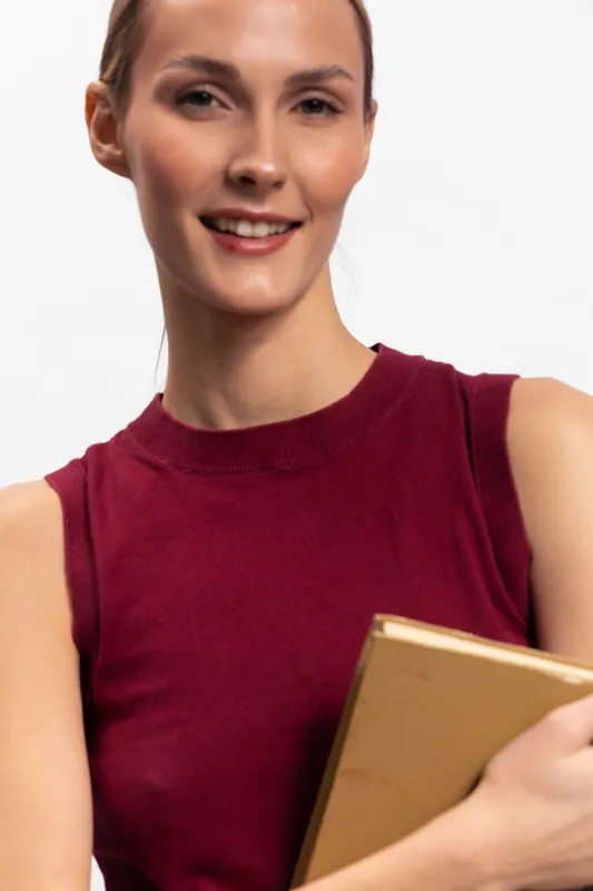 Smiling woman with brown hair wearing sleeveless red top holds a closed book against white background. Concept of education, knowledge, learning, and reading in minimalistic studio setting.