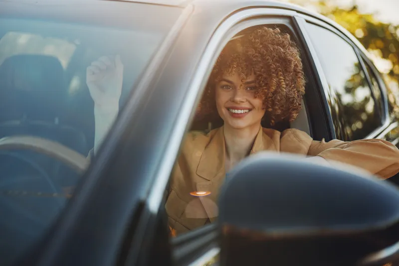 Smiling woman with curly hair sits inside a car, retro film color palette, casual lifestyle portrait, warm daytime light, candid moment behind the wheel, cheerful mood