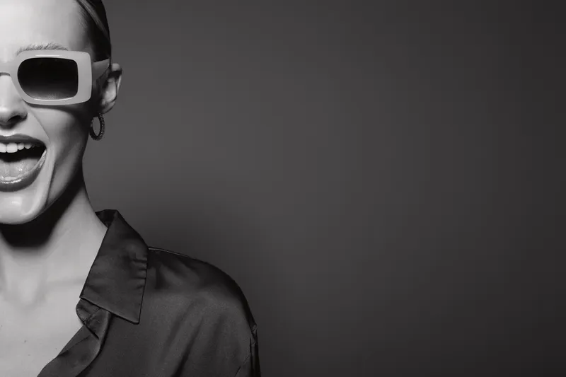 Close-up black and white photo of a stylish young white woman wearing sunglasses and a shirt, smiling widely with open mouth against dark background.