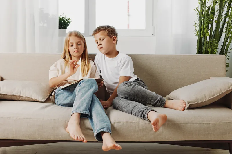 Two children sitting barefoot on a beige sofa in a bright room, focused on a tablet device. Casual clothing and natural light create a relaxed family and technology concept.