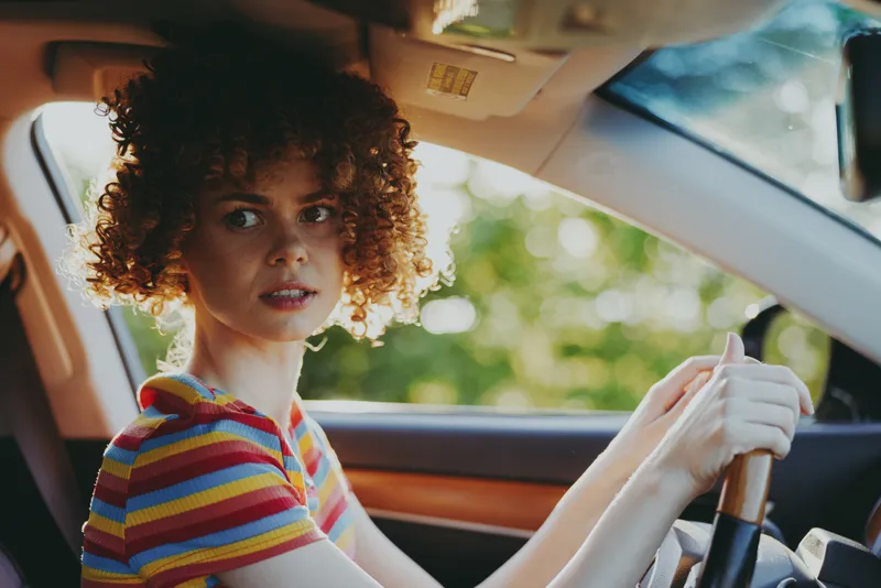 Young woman with curly hair drives a car, wearing a striped shirt, looking ahead with a focused expression, retro film color enhances warm interior light and nostalgic mood