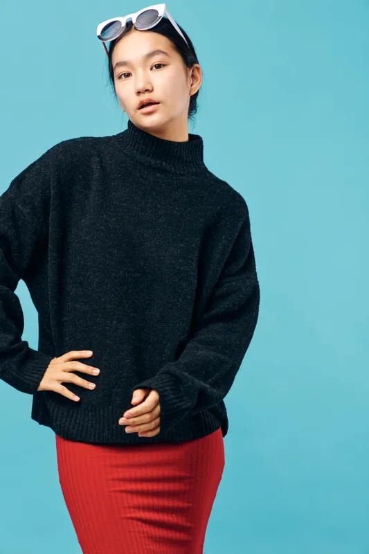 Young woman wearing black sweater and red skirt posing against blue background with sunglasses on head. Fashion, style, casual look with confident expression and modern outfit.