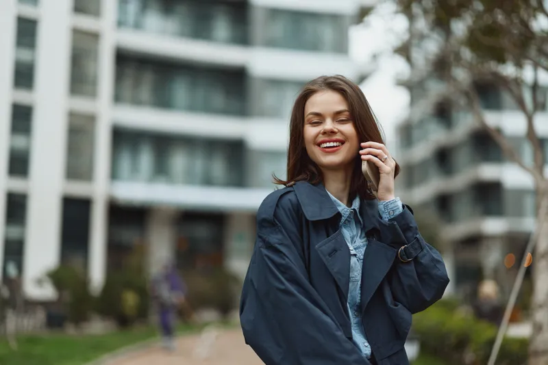 Young woman with long brunette hair smiles joyfully while standing outdoors in a casual blue jacket and denim shirt with an urban building background.