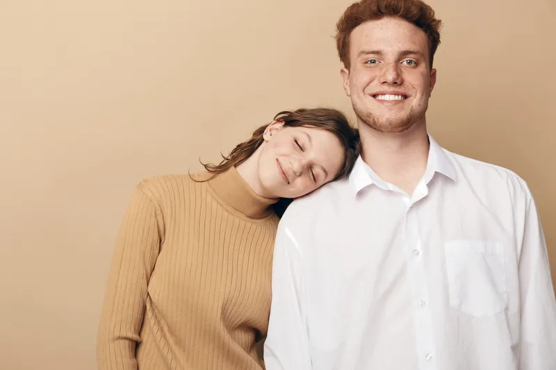 Smiling young man in white shirt stands next to woman in beige sweater who rests her head on his shoulder with closed eyes. Warm beige background creates cozy and intimate mood.