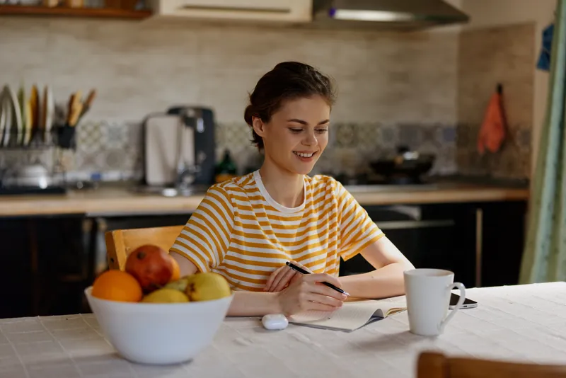 Young woman in a striped yellow shirt writes at a kitchen table, with a bowl of fruit nearby, focused and relaxed as she plans her day, in a sunlit room with warm morning light.