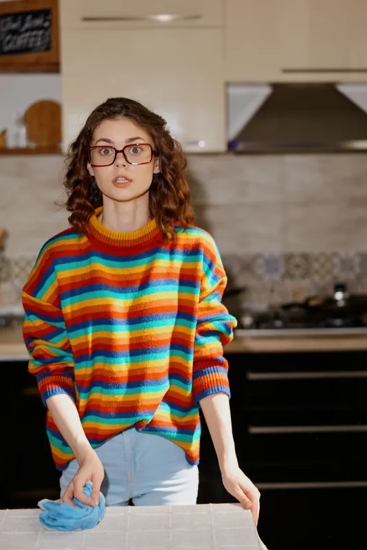 A young woman in a colorful striped sweater stands at a kitchen counter, glove on one hand, looking up with a surprised expression. A candid home lifestyle moment.