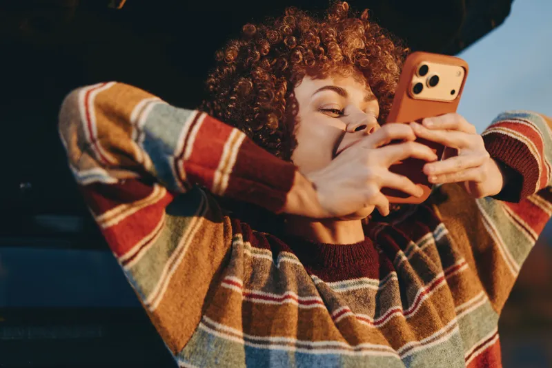 Lifestyle portrait of a woman with curly hair smiling while holding a smartphone to take a call indoors, warm lighting, casual striped sweater, relaxed pose, close view of hands