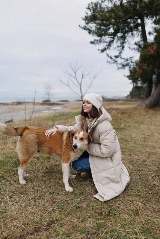 Woman kneels in a light coat with a beanie, pet dog by the shore in a chilly outdoor scene, calm sea and trees in the background, gentle moment