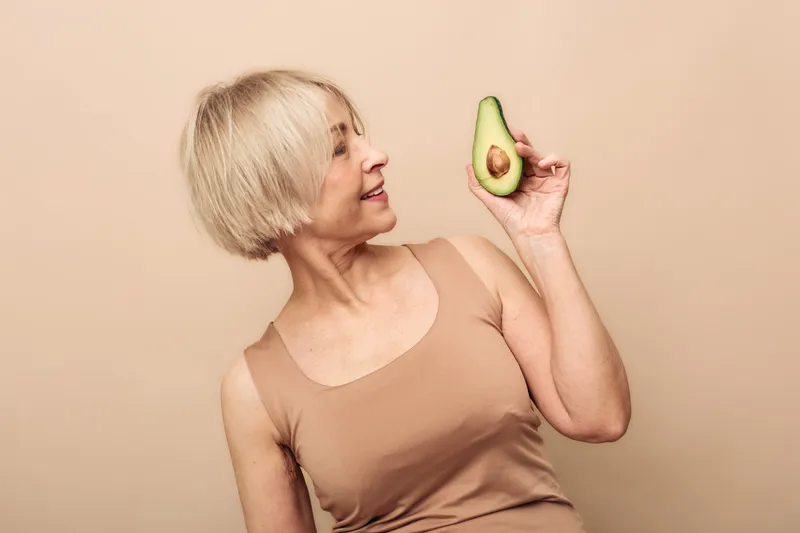 Mature woman with short blonde hair holds half avocado smiling on beige background. Concept of healthy lifestyle, nutrition, natural food, and positive wellbeing for older adults.