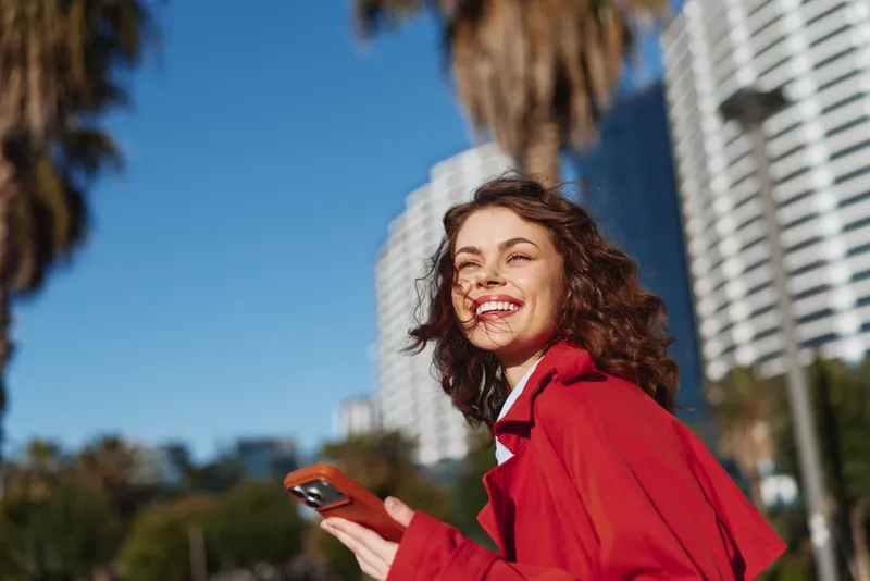 Young woman in a red jacket laughs outdoors, holding a phone in a sunny urban setting with palm trees and tall buildings behind, radiating confidence.