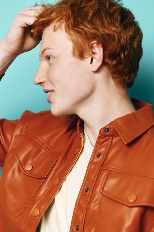 Redhead man in profile with hand in hair wearing brown leather jacket and white shirt standing against blue background with thoughtful expression.