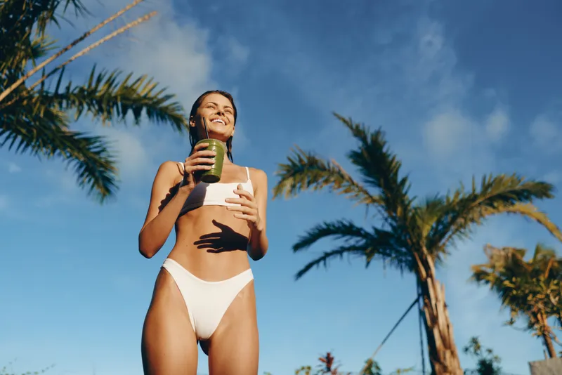 Smiling woman in a white bikini enjoying a smoothie outdoors under a sunny sky among palm trees. Lifestyle imagery focused on wellness, self care, and outdoor vitality.