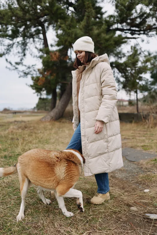 Woman in a warm puffer coat and beanie outdoors with a dog on a leash in a park, calm natural setting with trees and grass, candid moment of companionship and exploration