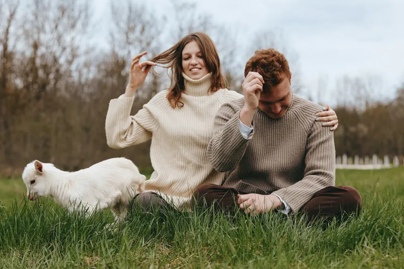 Happy couple sits on grass in cozy sweaters, enjoying a relaxed moment together in a park, with a small white pet nearby, creating a warm lifestyle scene of comfort and connection.