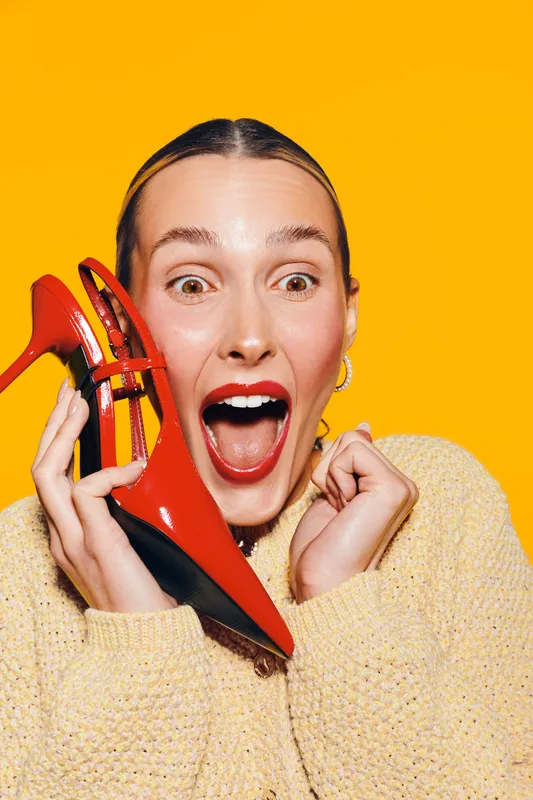 Excited woman with red lipstick holds red high heel shoe near face on yellow background. Concept of fashion, style, shopping, and happiness with bright studio lighting.