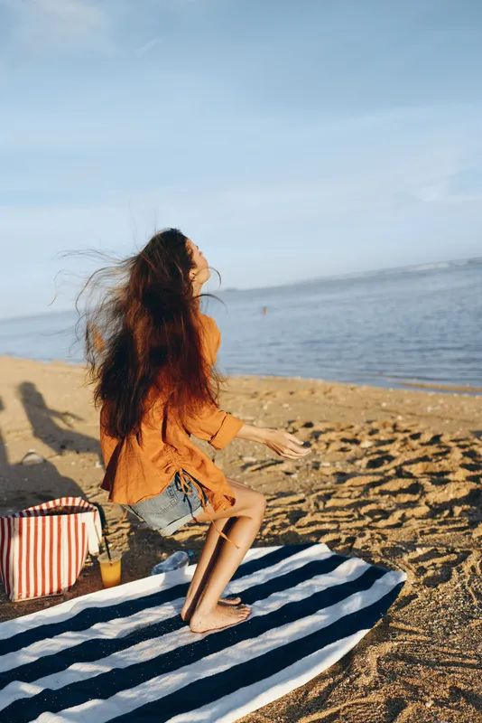 woman beach ocean sunlight relaxation summer sitting blanket striped serenity, a woman in orange dress sits on a striped blanket on sandy shore near the sea, red hair flowing in wind, ocean waves and
