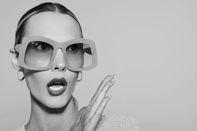 Surprised young Caucasian woman with fair skin wears oversized sunglasses and hoop earrings, posing with open mouth and raised hand. Fashion and expression concept in studio.