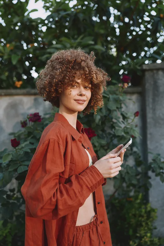 Young woman with curly hair in an orange outfit stands outdoors, looking at her phone amid blooming roses and a stone wall, exuding calm concentration and urban garden mood