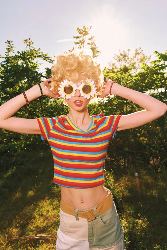 A woman in a striped top poses outdoors in bright sunlight, wearing large daisy sunglasses. Retro color grading enhances a playful, carefree vibe and summery mood.