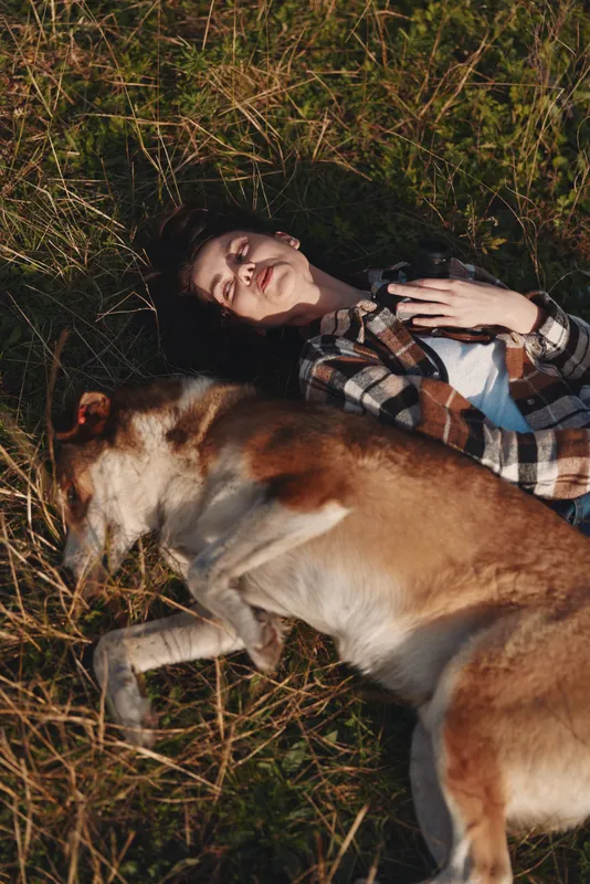 Dog and woman resting together in a sunlit meadow, smiling as they lie among tall grass. Casual plaid shirt, relaxed pose, warm connection and peaceful outdoor moment for lifestyle, friendship and
