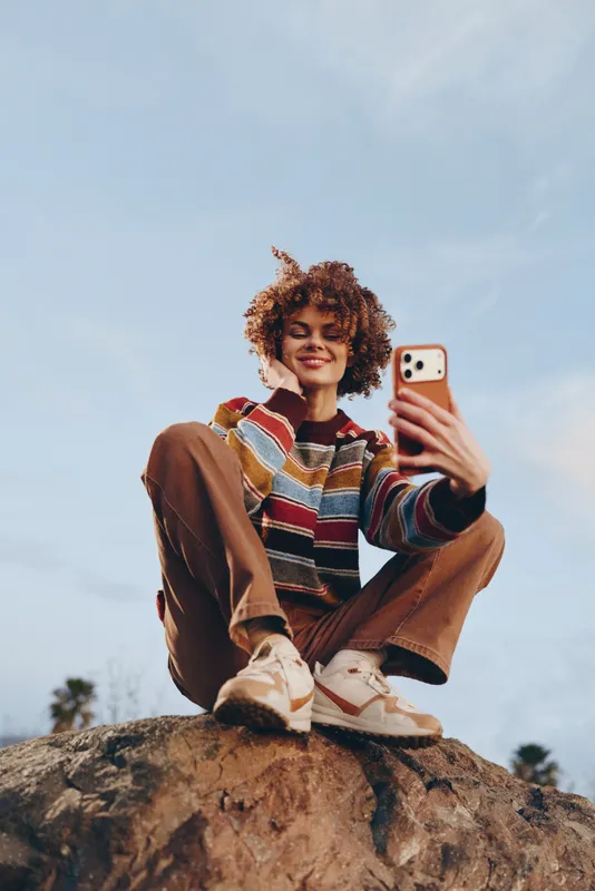 Woman selfie with smartphone outdoor, casual fashion lifestyle portrait, sitting on rock beneath clear sky, curly hair smiling, wearing colorful sweater and cozy shoes, sunlit summer leisure