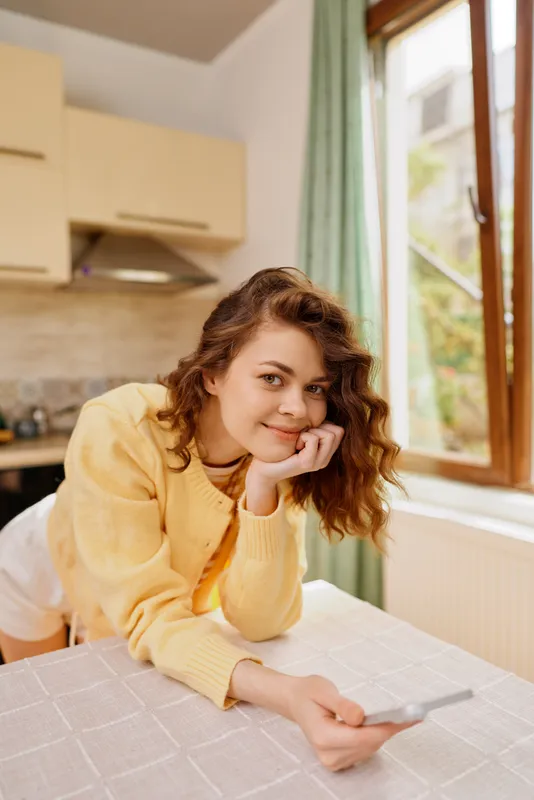 A woman in a yellow cardigan lounges at home, resting her chin on her hand while holding a card. The bright, cozy room conveys a casual, relaxed lifestyle and everyday selfcare.