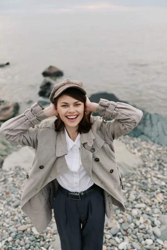 Smiling young woman in warm knitted cardigan and hat stands on rocky beach by calm sea, expressing happiness and relaxed mood in casual autumn outfit outdoors.