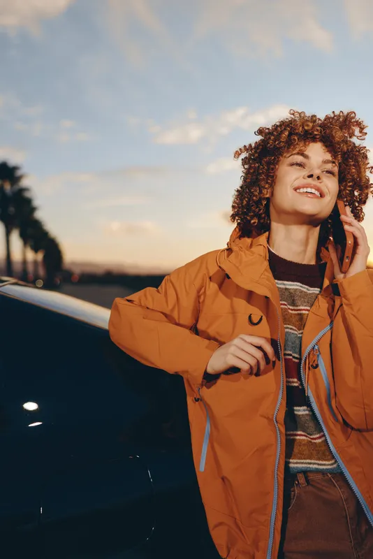 Woman lifestyle outdoors, smiling in warm sunset light, holding phone to ear, standing by car, brown jacket, curly hair, relaxed pose, evening road background, soft breeze, trees nearby