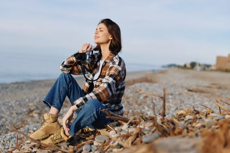 Woman photographer sits on a pebbled beach with her camera, wearing a plaid shirt and denim jeans, enjoying the coast in a calm outdoor moment, sunny weather.