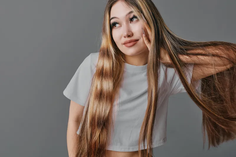 Young woman with long straight hair posing, wearing casual white t shirt against neutral background, looking away with thoughtful expression and hand near face.