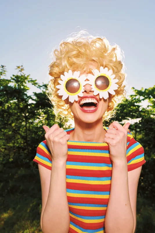 Playful young woman wearing sunflower sunglasses and a striped tee grins wide in a sunlit park, embracing a retro film color mood and carefree summer energy.