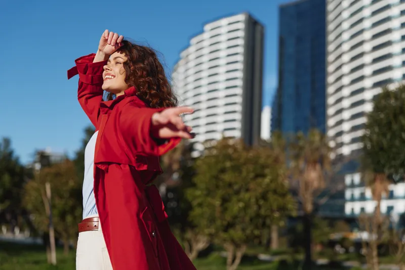 Adult woman with curly brunette hair wears a red coat in an outdoor city park, striking a dynamic pose with an outstretched arm under a bright blue sky. She radiates energy. Today.