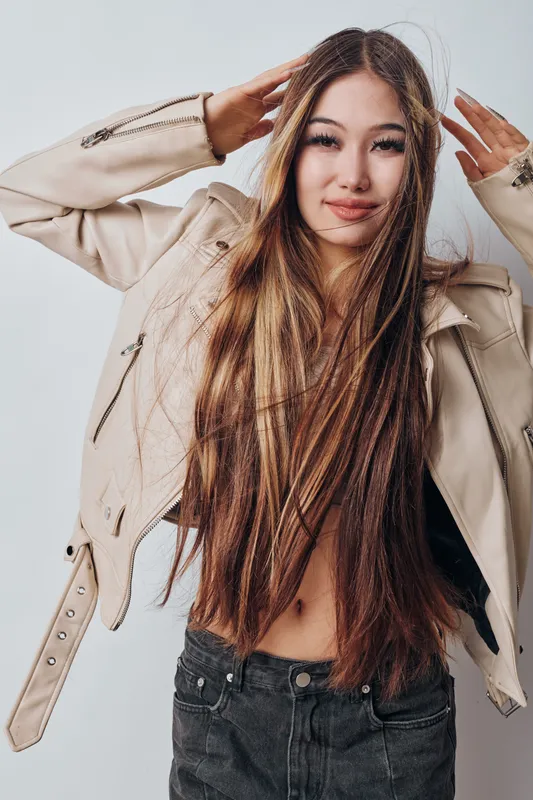 Young woman with long straight hair wearing light jacket and dark pants posing with hands near head against white background looking at camera smiling.