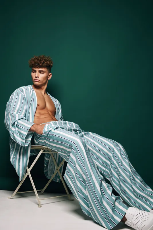 Young man sitting casually on chair wearing striped loose shirt and pants looking sideways with thoughtful expression. Stylish modern fashion portrait with simple green background in studio.