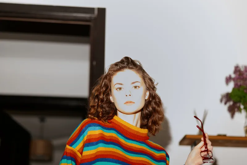 A young woman wearing a striped colorful sweater stands indoors, looking directly at the camera with a calm, thoughtful expression, capturing a candid home lifestyle moment.