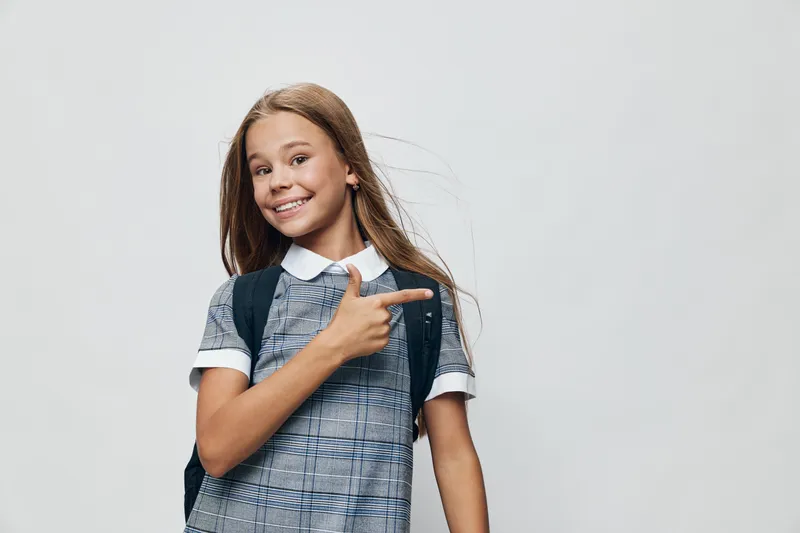 Girl smiling and pointing finger to side in casual outfit with backpack on gray background. Youth, happiness and gesture for positive expression and school or lifestyle concept.