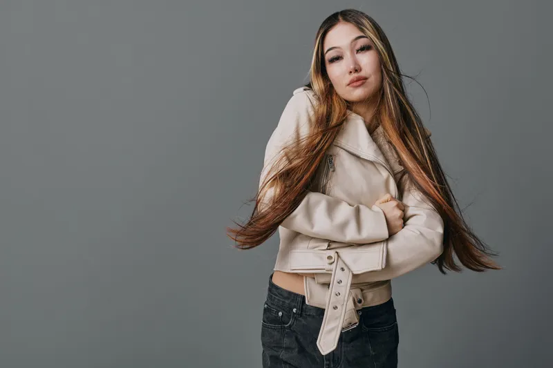 Young woman with long hair wearing a beige jacket and black jeans posing confidently on gray background, fashion portrait with natural look and casual style indoors.