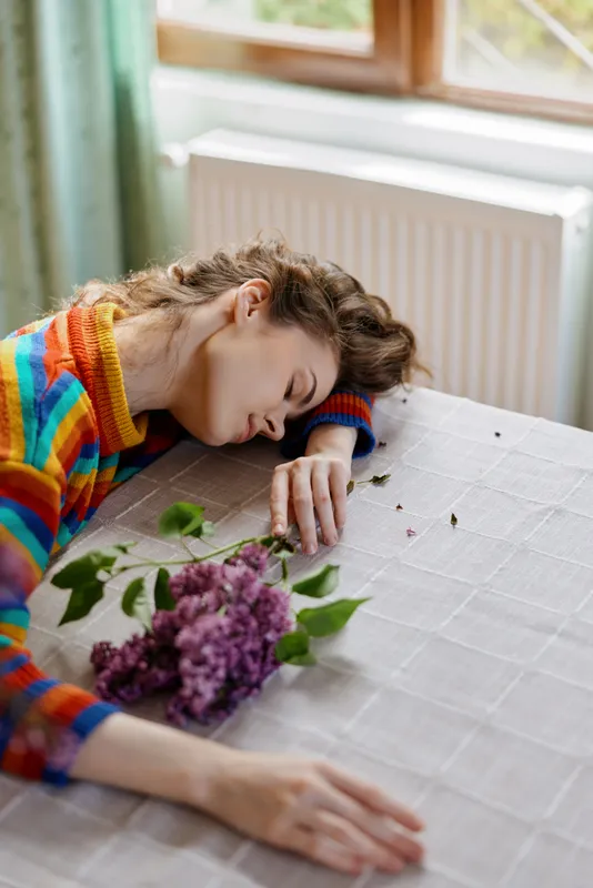 A sleeping woman rests her head on a light dining table indoors, a colorful striped sweater draped over her and a bouquet of lilac flowers nearby, conveying quiet fatigue and tenderness